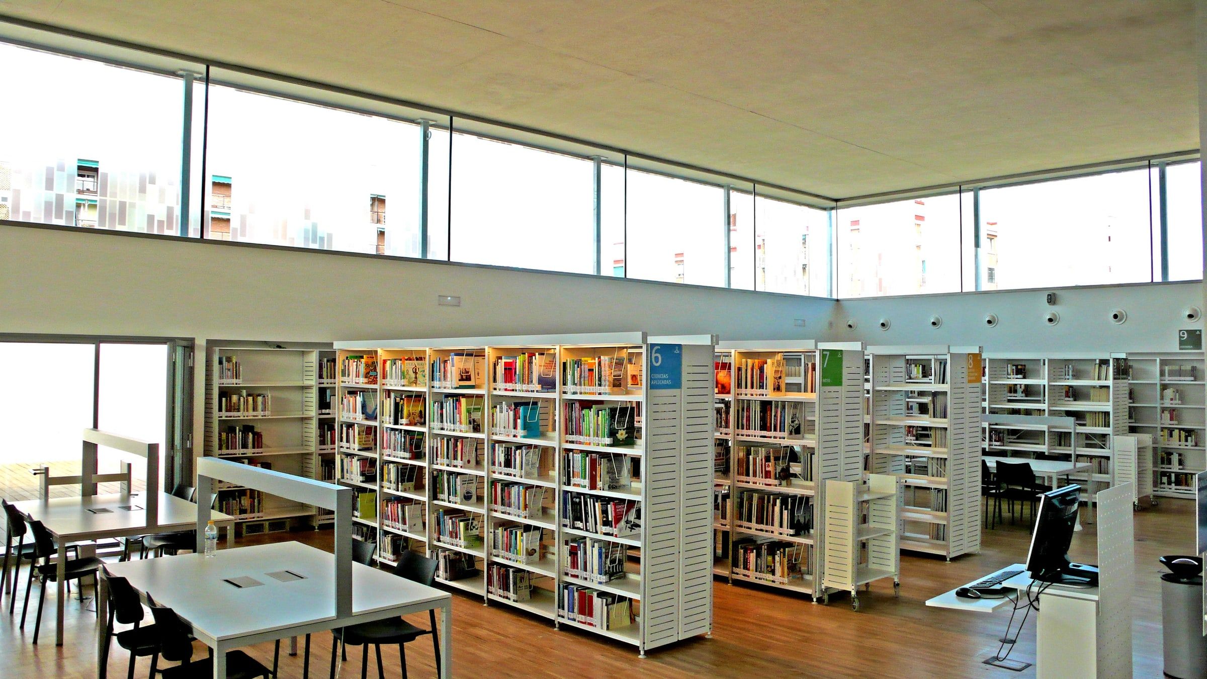 Sala de lectura y préstamo de una biblioteca pública municipal, con estanterías de libros, mesas de estudio y grandes ventanales