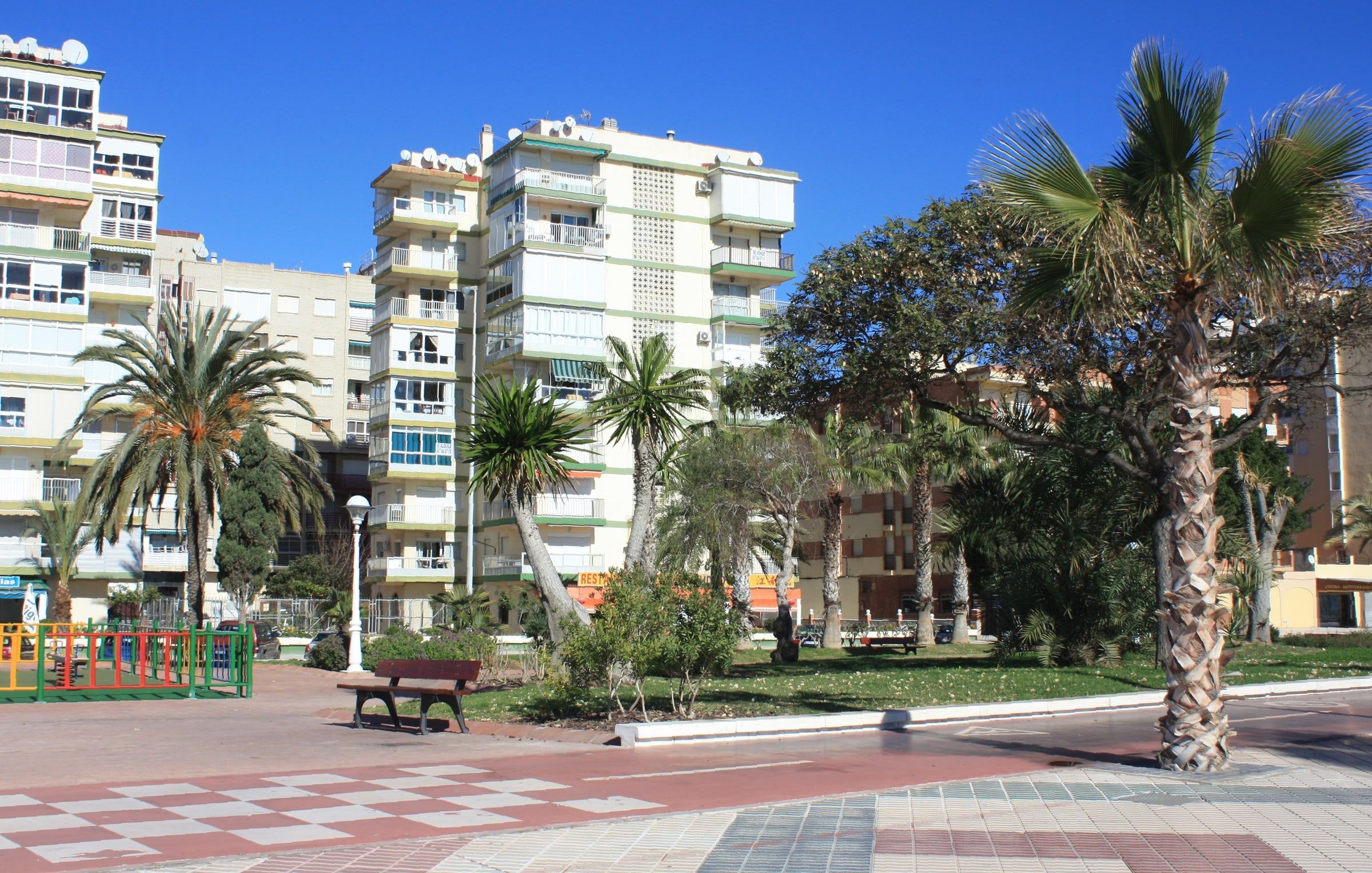 Bloques de apartamentos frente al paseo marítimo de Levante de Torre del Mar, con palmeras, jardines y bancos bajo el cielo azul