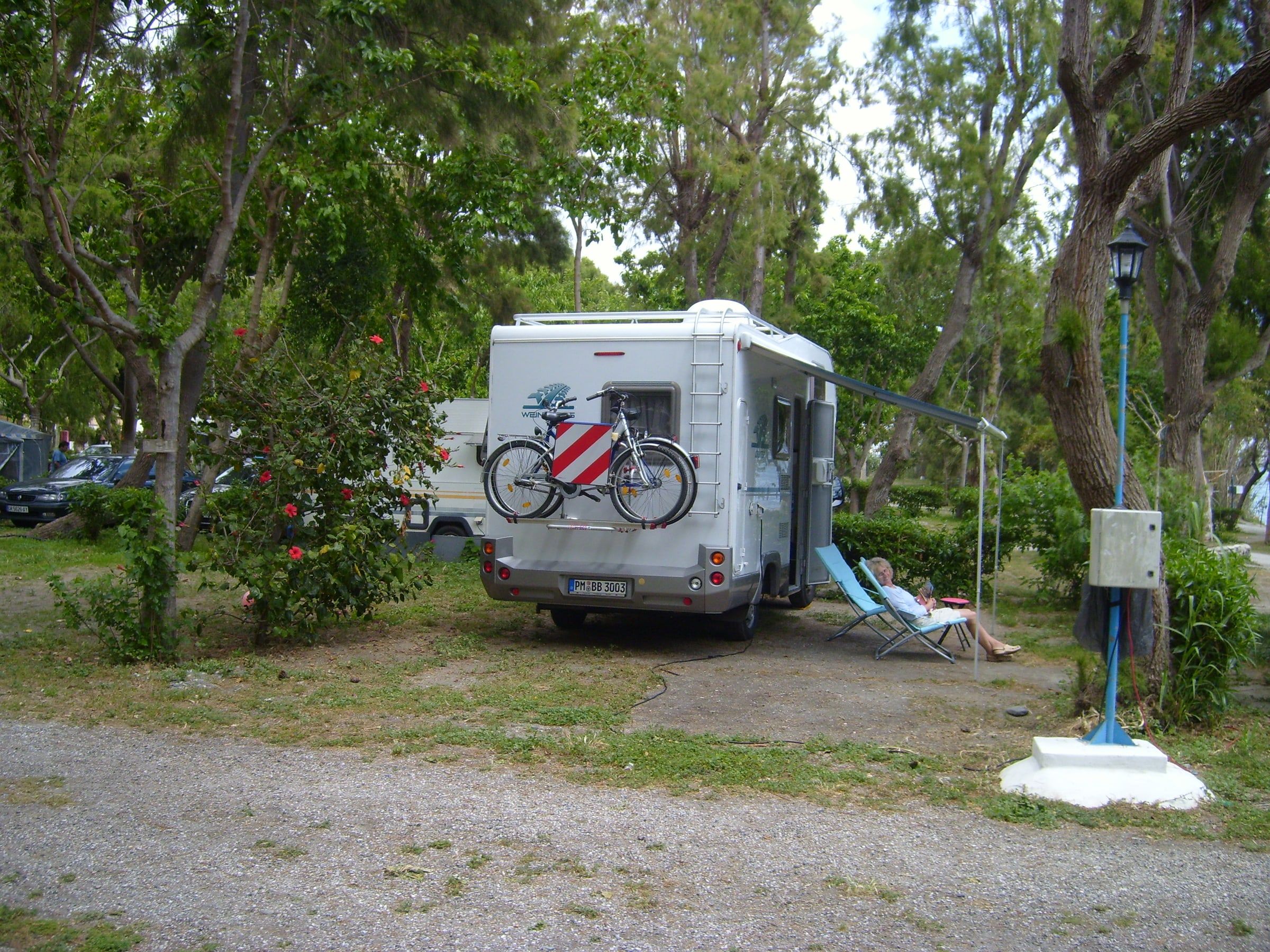 Autocaravana con bicicletas y toldo aparcada en una parcela de un camping de la costa, a la sombra de árboles y adelfas