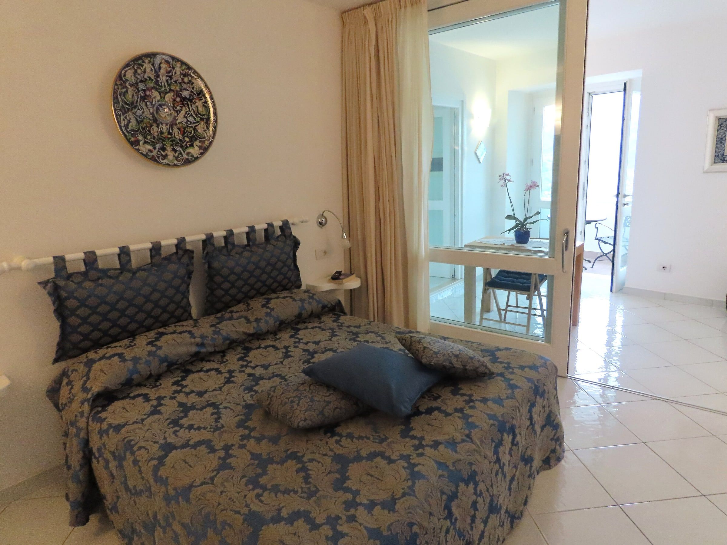 Habitación doble sencilla de un pequeño alojamiento, con colcha azul estampada, suelo de baldosa clara y una ventana luminosa
