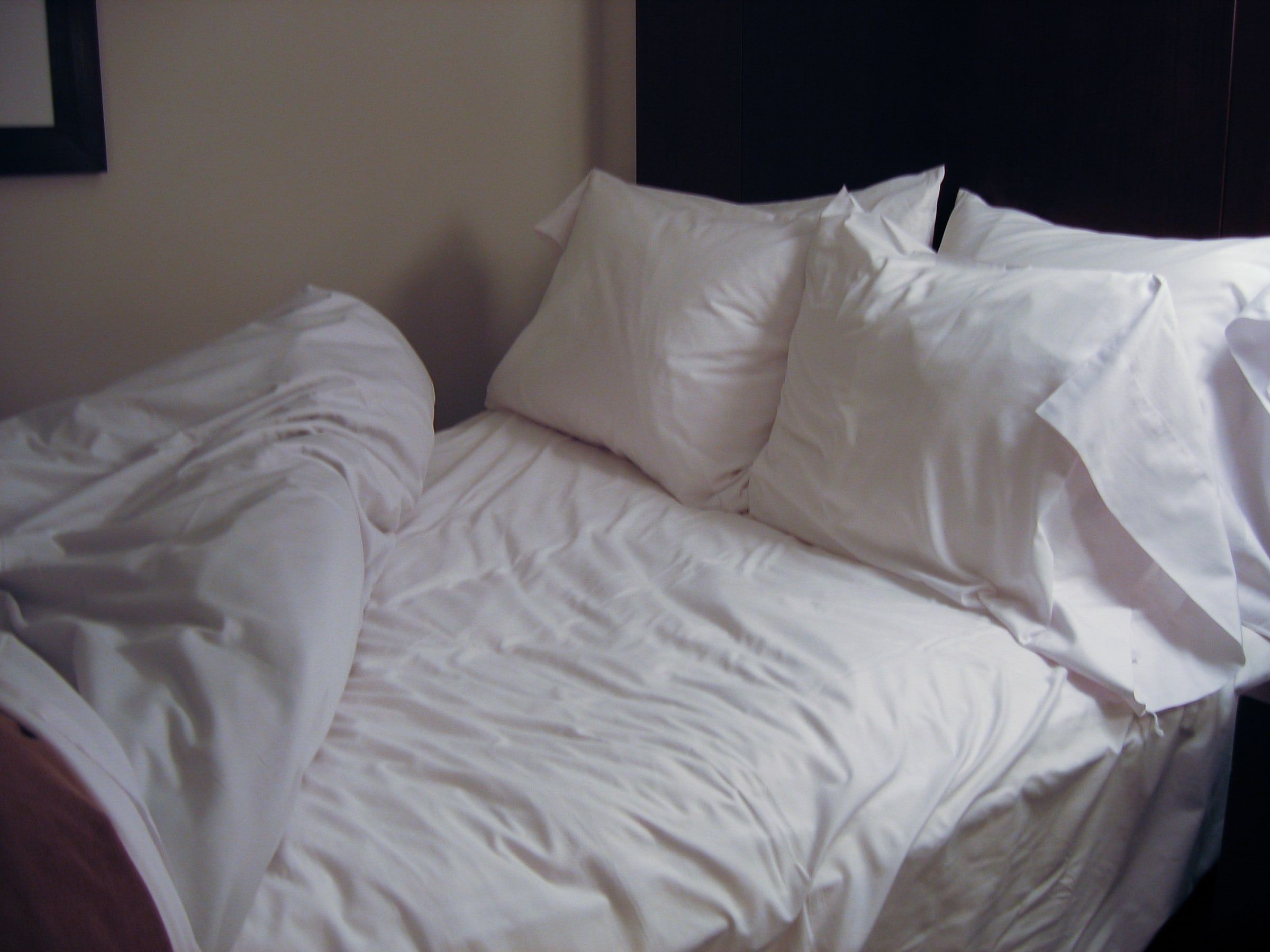 Cama de matrimonio con sábanas blancas recién usadas y cuatro almohadas mullidas, en un dormitorio con luz suave