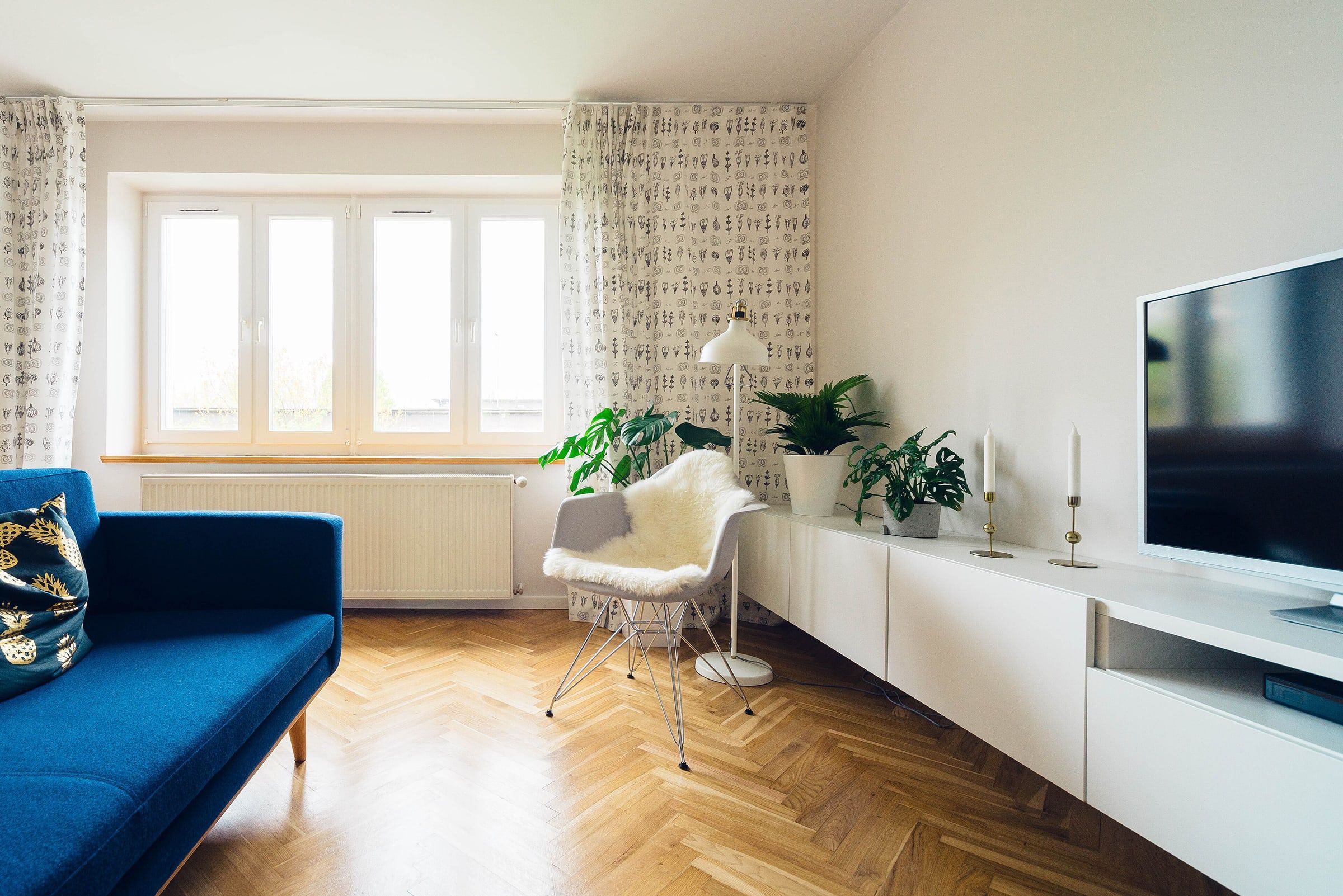 Salón luminoso con sofá azul, butaca blanca, mueble bajo, plantas y suelo de madera en espiga junto a la ventana