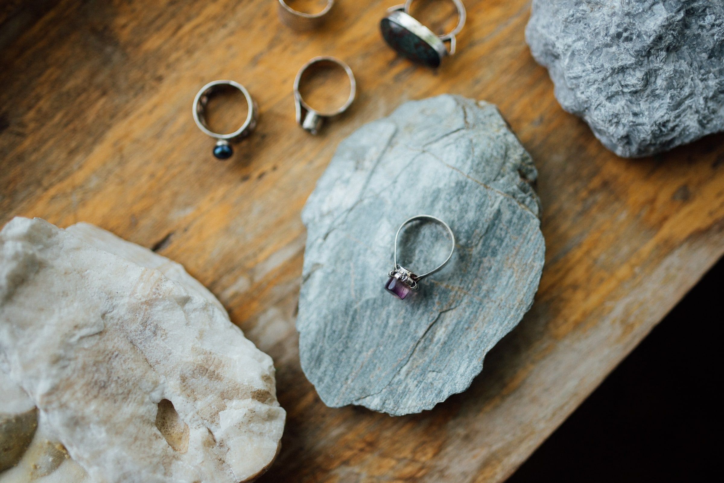 Anillos de plata, uno de ellos con una piedra morada, colocados sobre piedras lisas y una tabla de madera a la luz del día