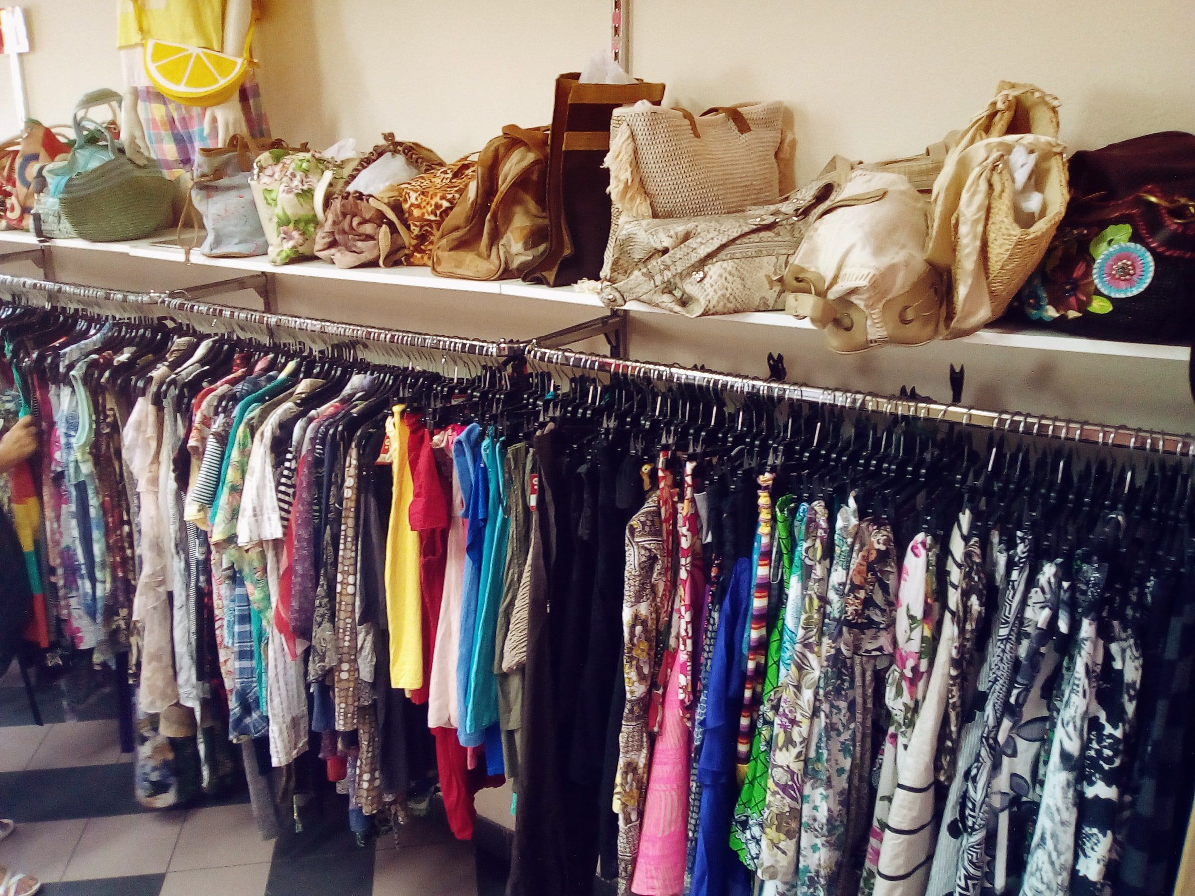 Barra de una tienda de ropa con prendas de colores y estampados colgadas en fila y varios bolsos en el estante de arriba