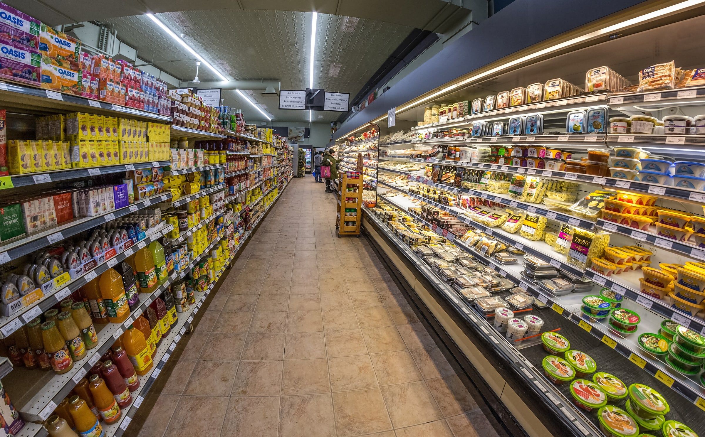 Pasillo de un supermercado con estanterías de conservas y bebidas a un lado y vitrinas refrigeradas al otro