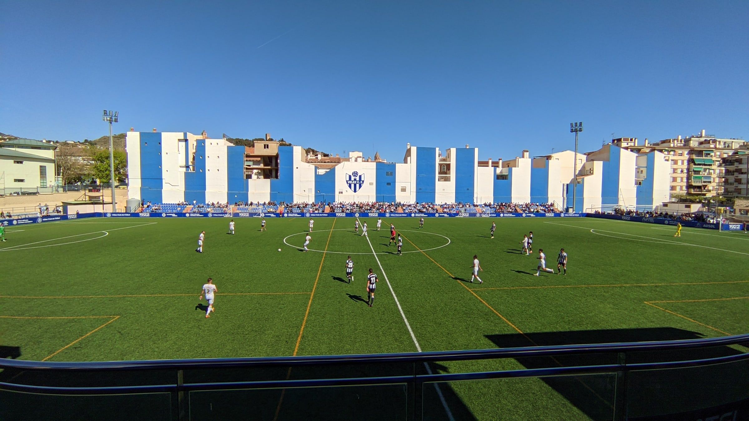 Partido de fútbol en el estadio Vivar Téllez de Vélez-Málaga, con el césped artificial y la grada pintada de franjas azules y blancas
