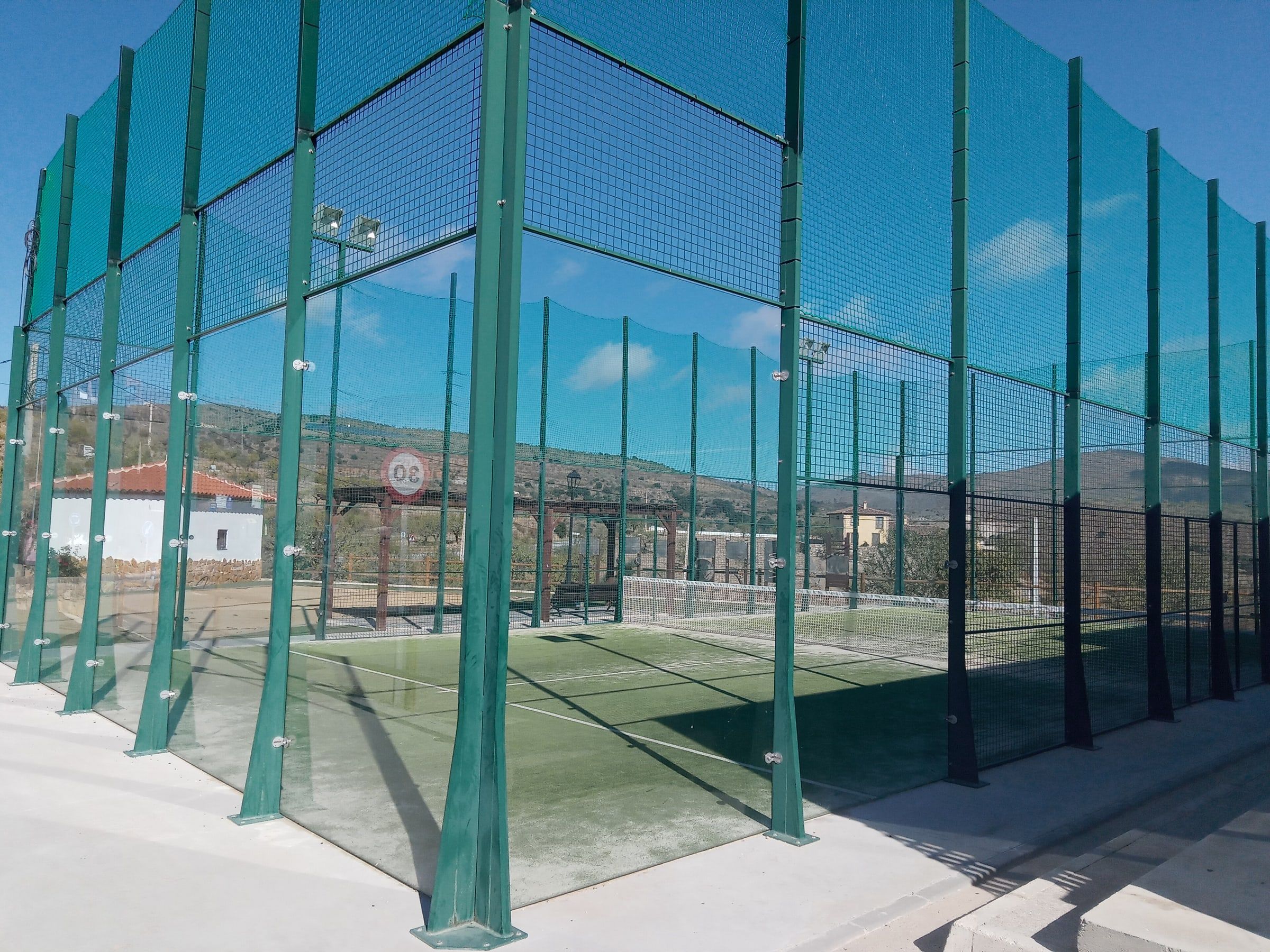 Pista de pádel al aire libre con paredes de cristal, malla metálica verde y césped artificial, en un pueblo de Andalucía bajo el cielo azul