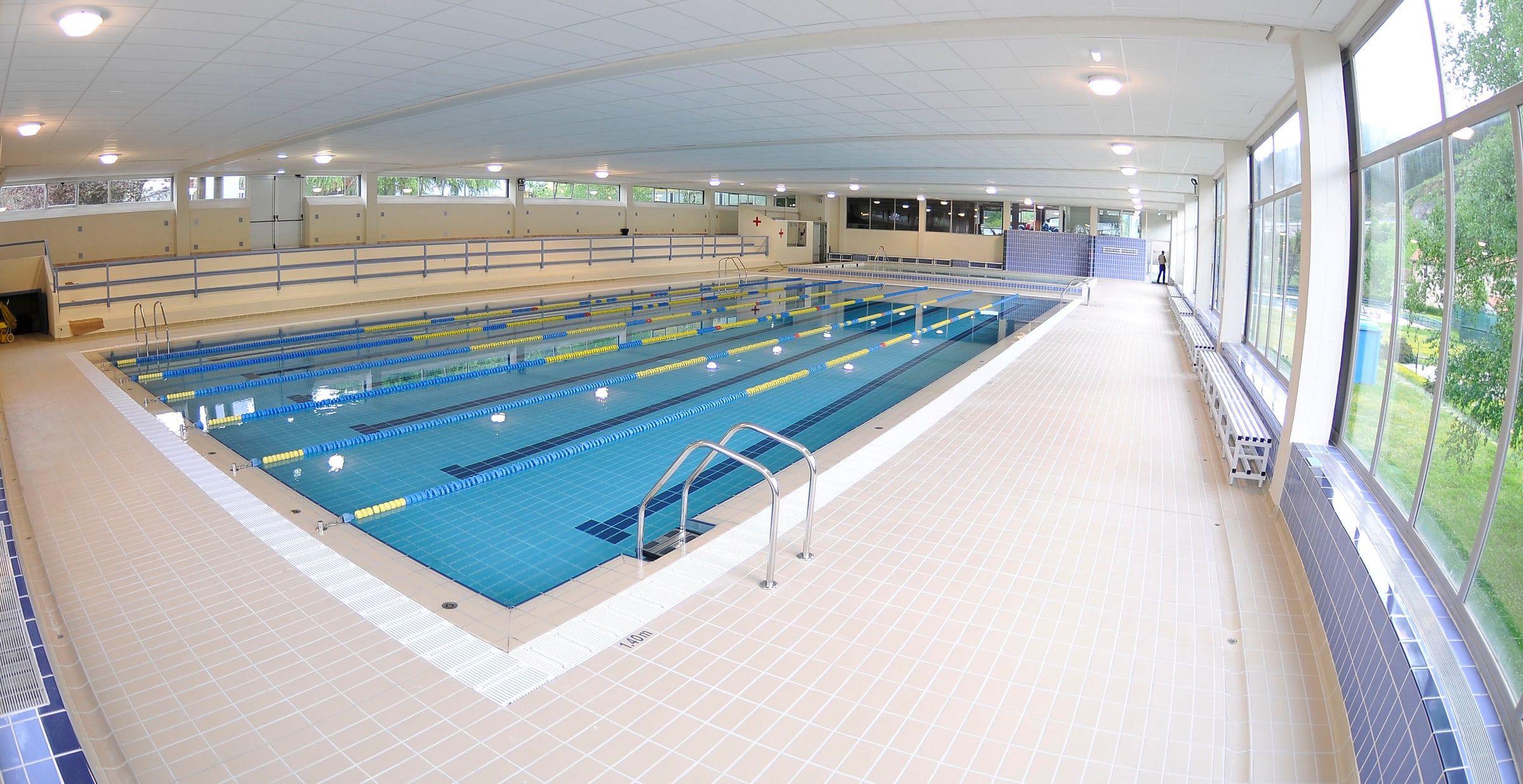 Piscina cubierta municipal vacía, con calles separadas por corcheras, escalerilla metálica y grandes ventanales a un lado