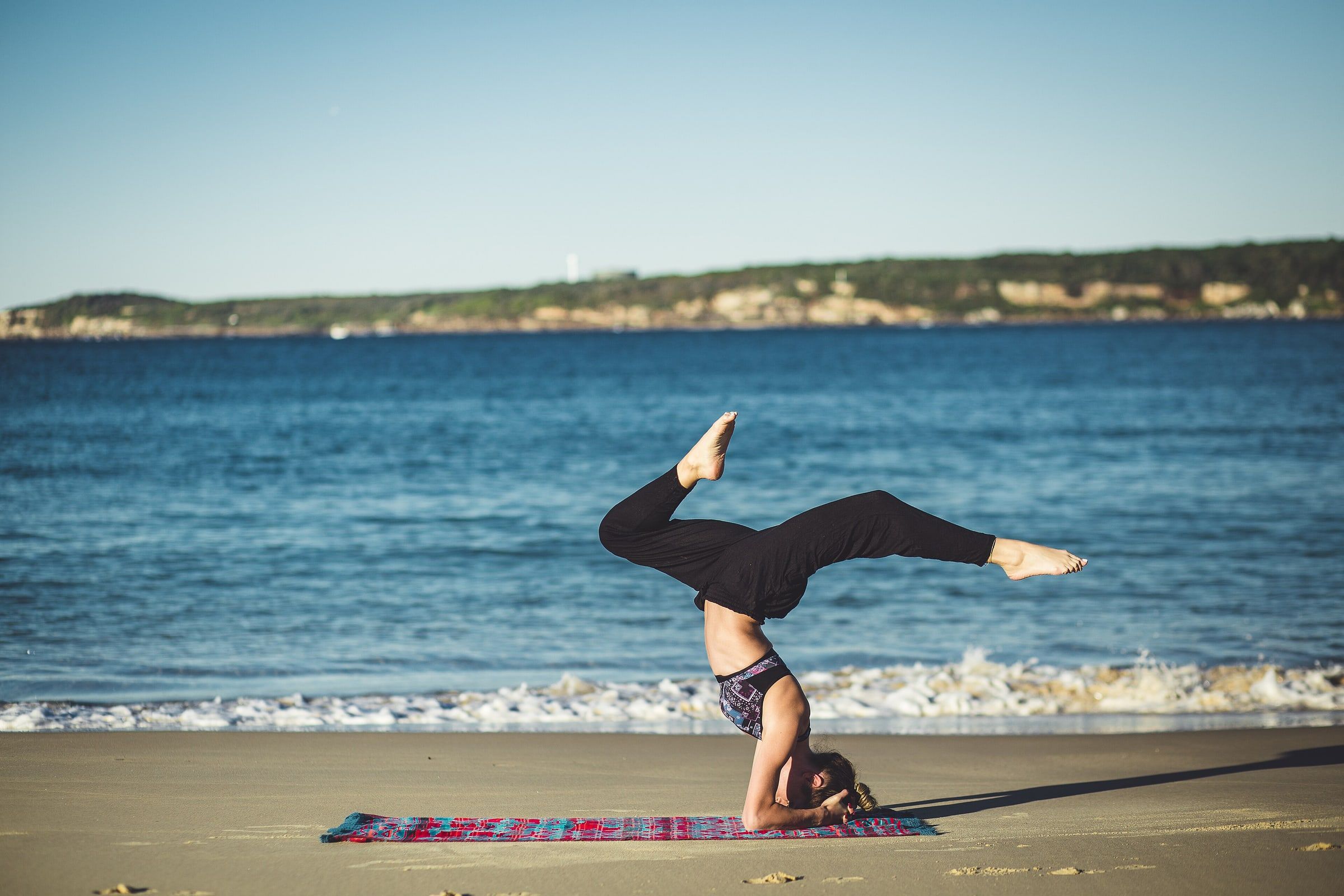 Mujer haciendo una postura de yoga invertida sobre una esterilla en la orilla de la playa, con el mar azul y la costa al fondo