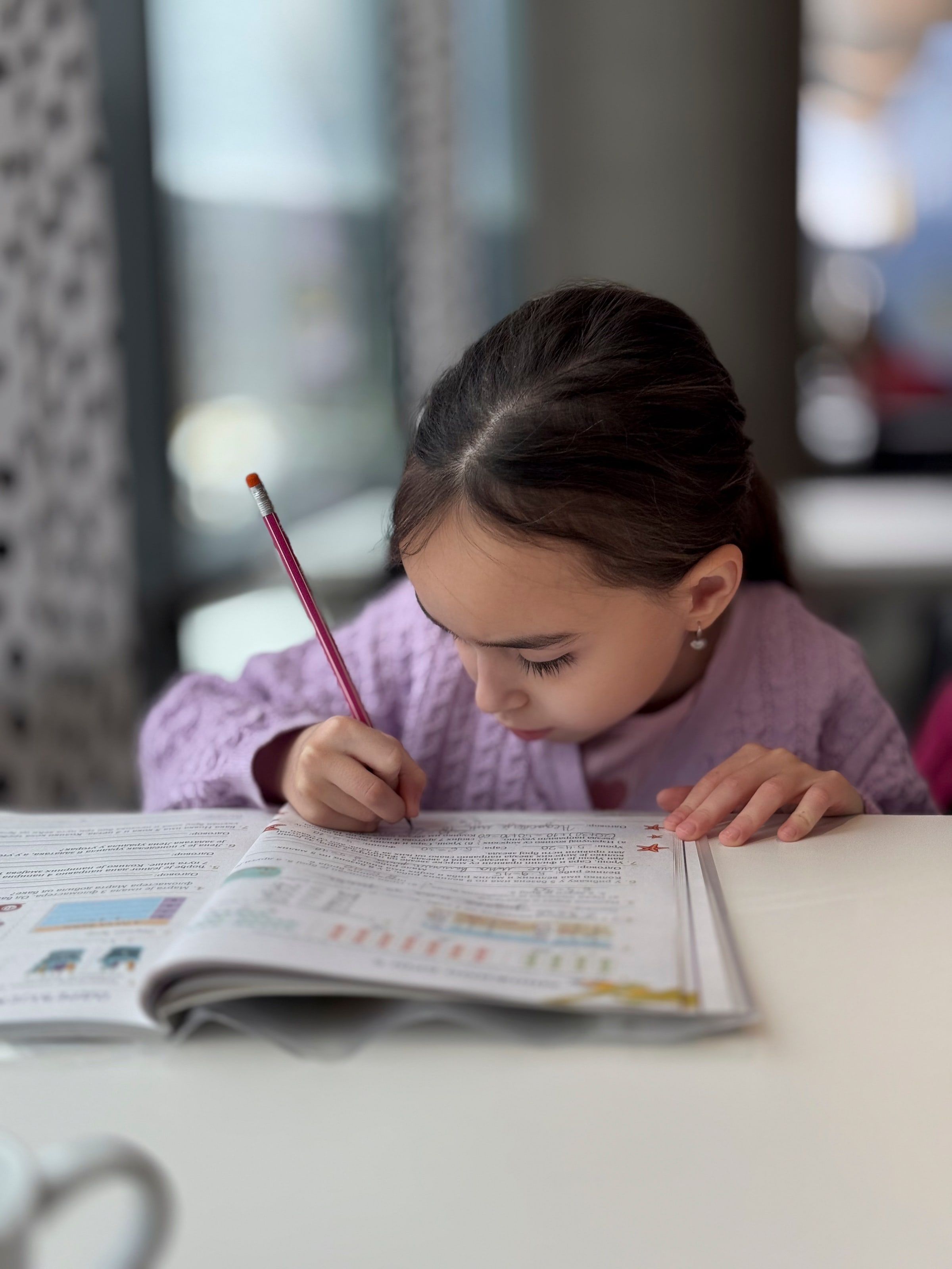 Niña haciendo los deberes en una mesa, escribiendo con lápiz en un cuaderno de ejercicios abierto