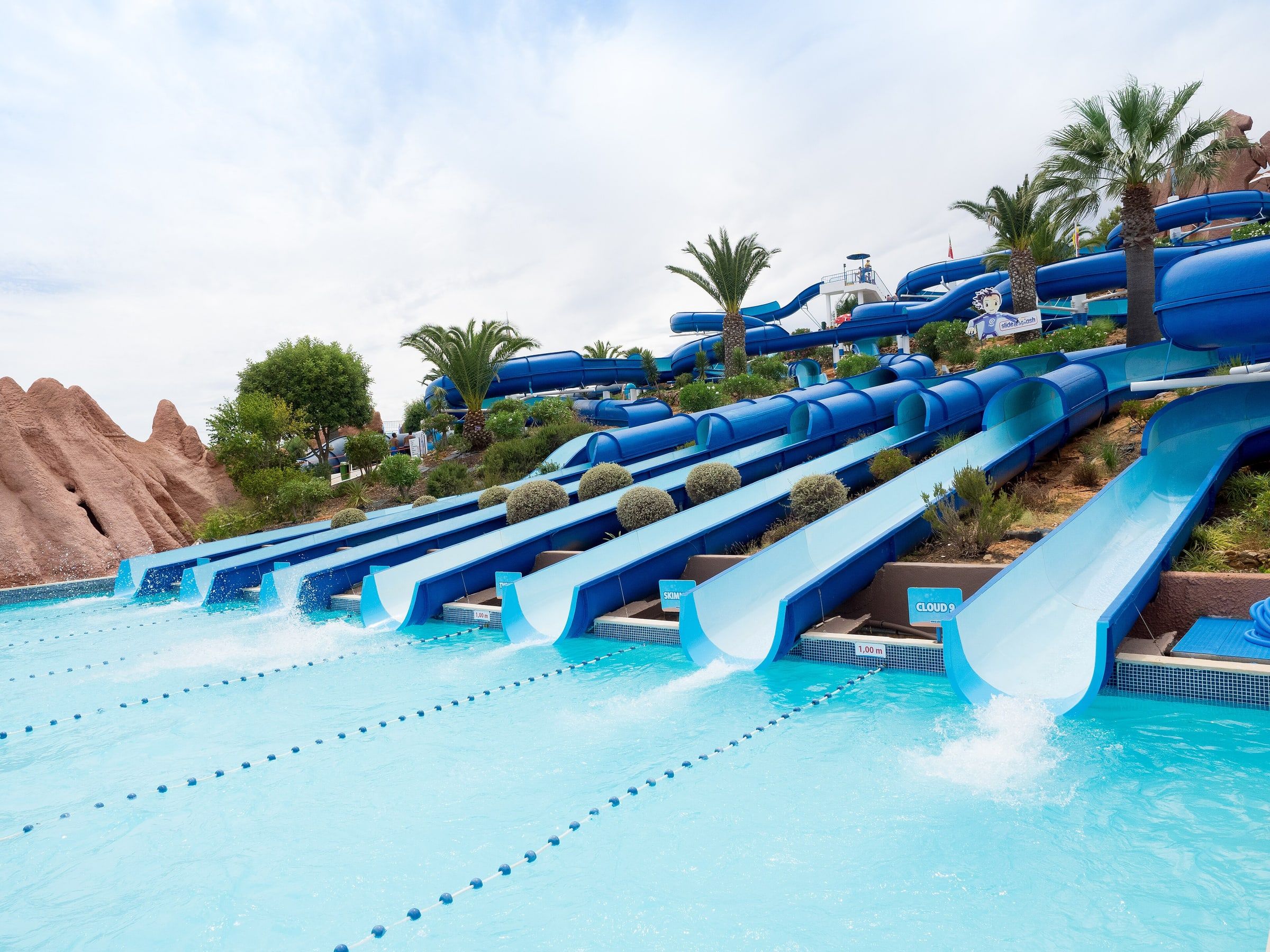 Toboganes azules de un parque acuático que bajan en paralelo hasta una piscina de agua turquesa, entre palmeras y rocas artificiales