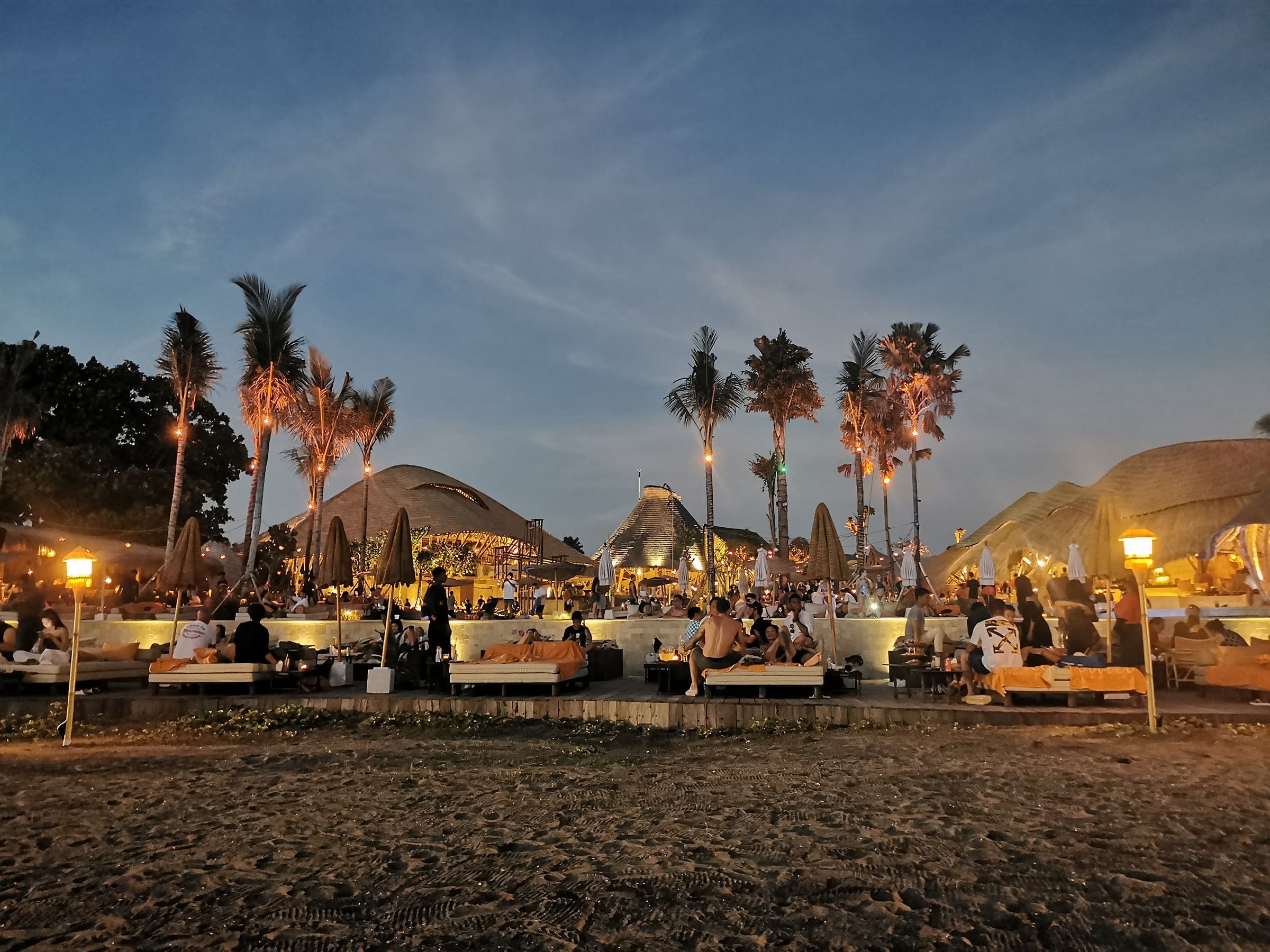 Club de playa al anochecer visto desde la arena, con farolillos encendidos, palmeras, sombrillas y gente en las hamacas y mesas