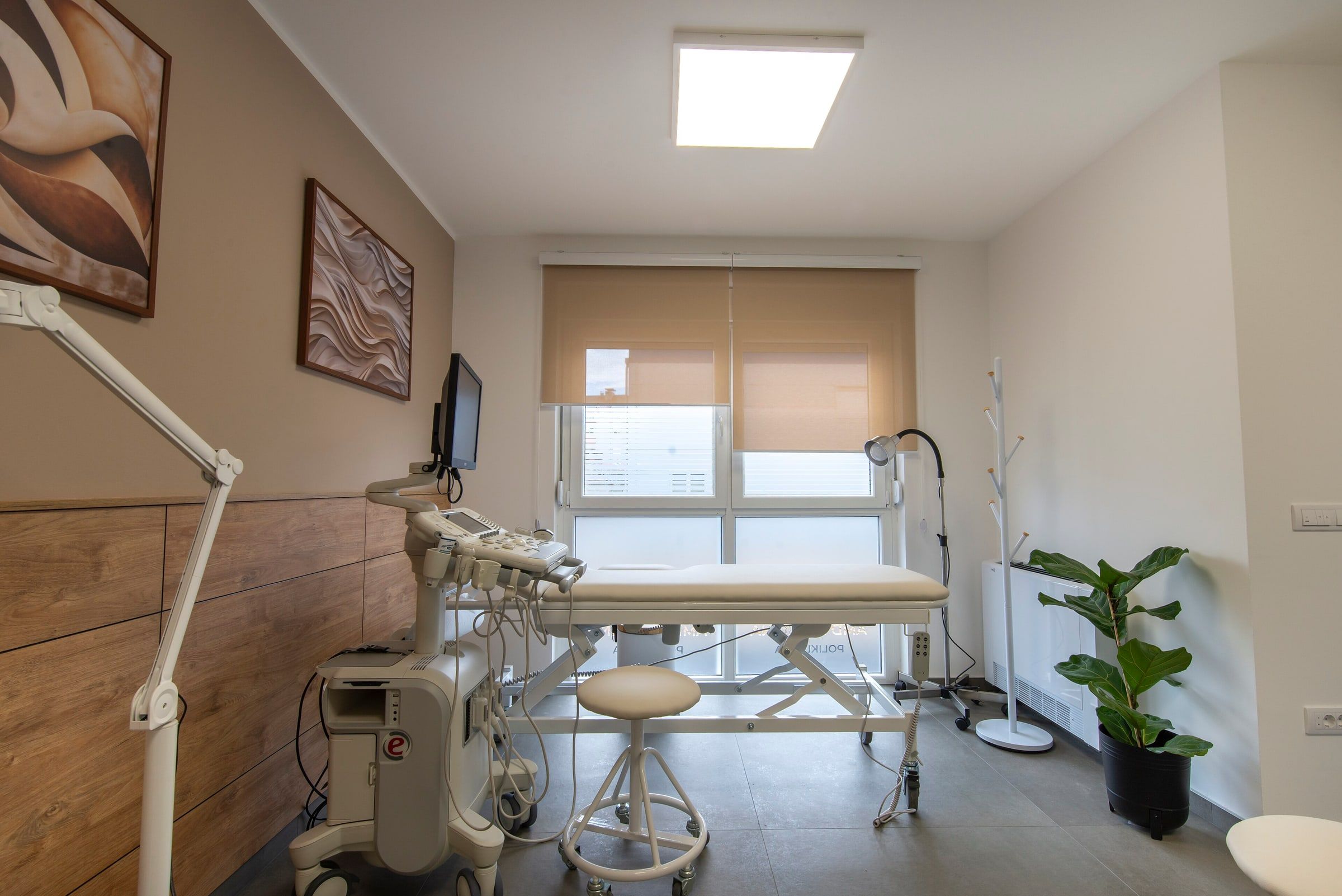 Sala de consulta de una clínica moderna con camilla, lámpara de exploración, aparatos médicos y una planta junto a la ventana
