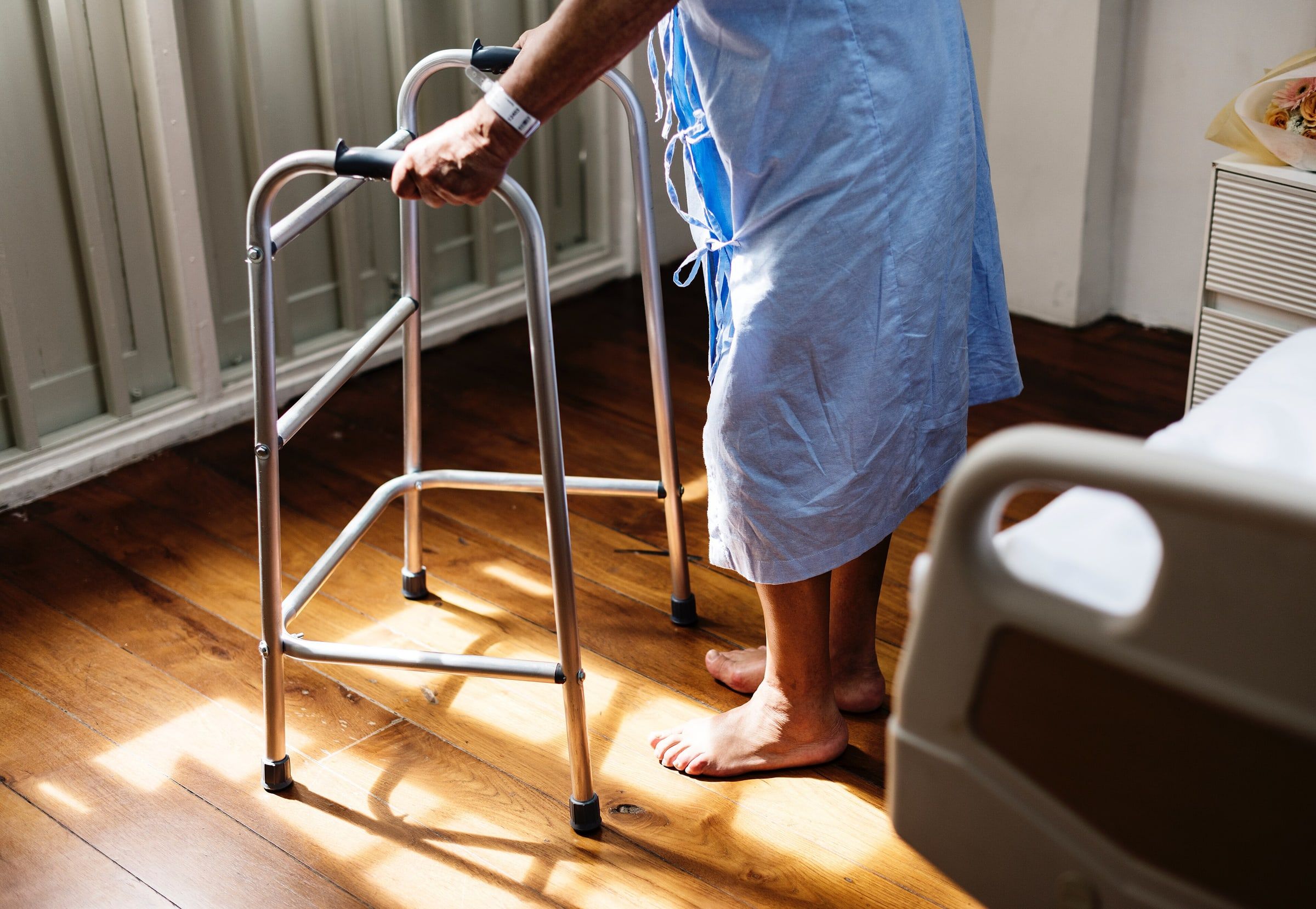 Paciente con bata de hospital apoyada en un andador metálico junto a la cama, vista de cintura para abajo sobre suelo de madera