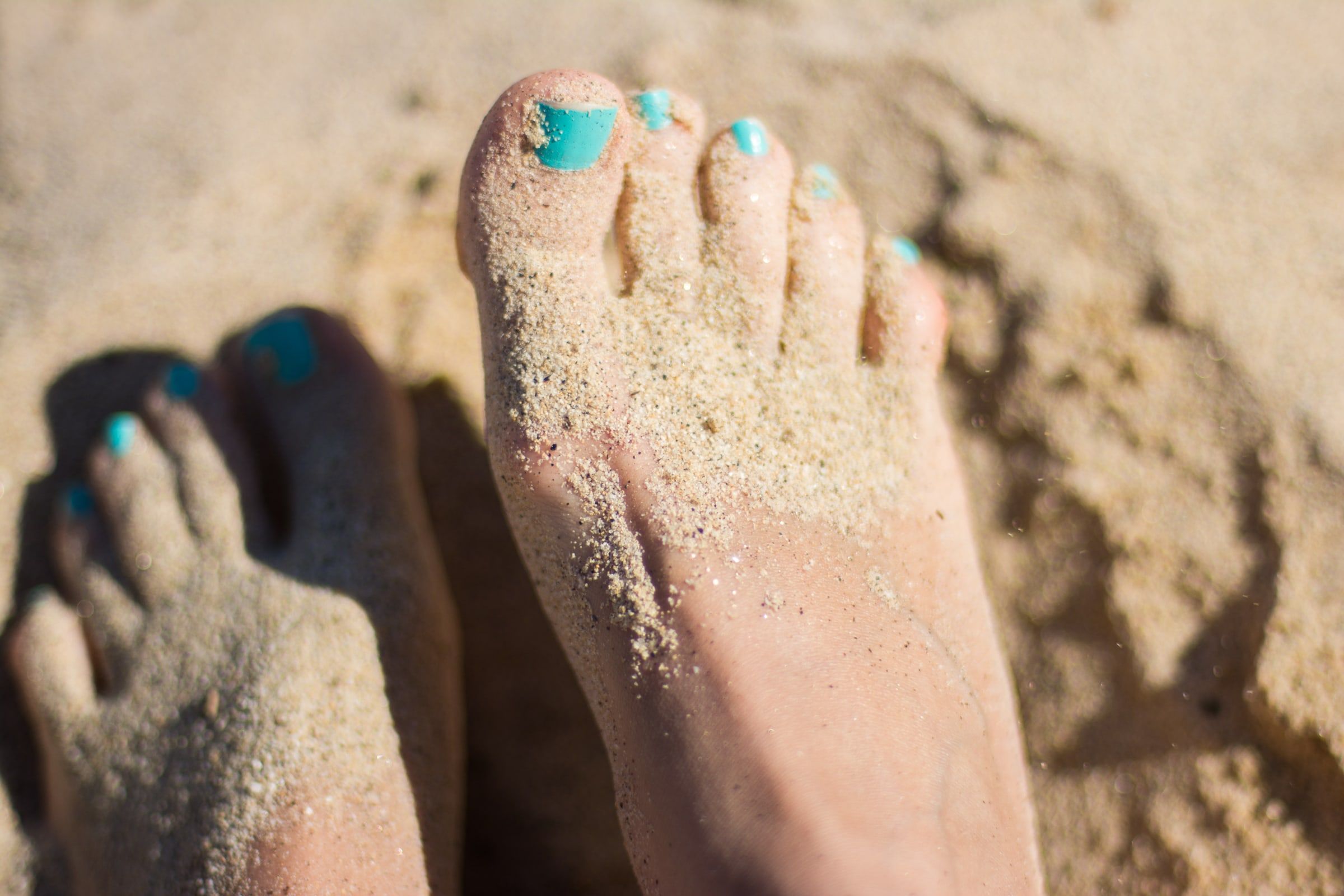 Pies descalzos cubiertos de arena en la playa, vistos de cerca, con las uñas pintadas de color turquesa