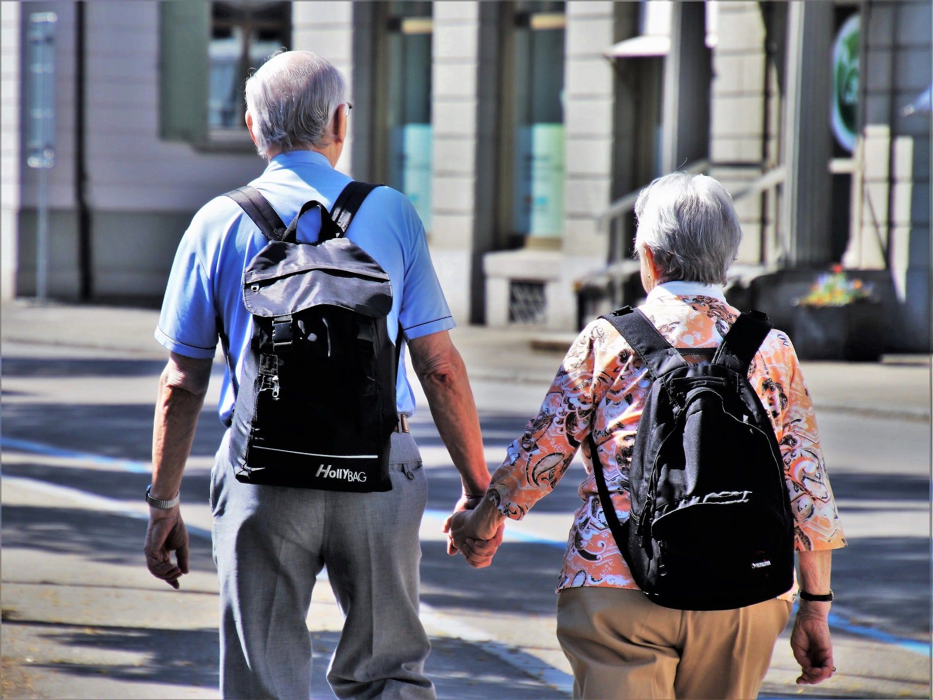 Pareja de personas mayores paseando de la mano por la calle con mochilas a la espalda, vistas por detrás