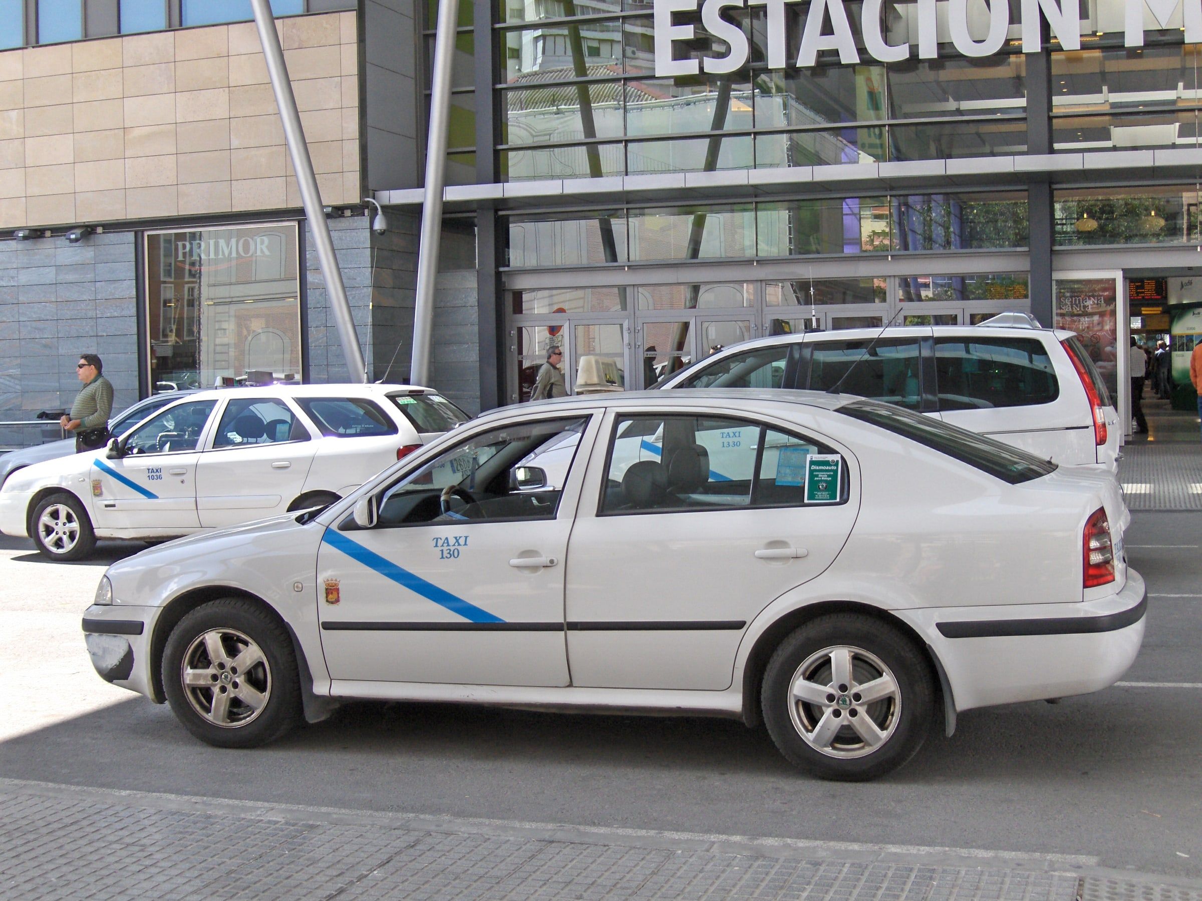 Taxis blancos con la franja azul de Málaga esperando en la parada a la salida de una estación, uno detrás de otro